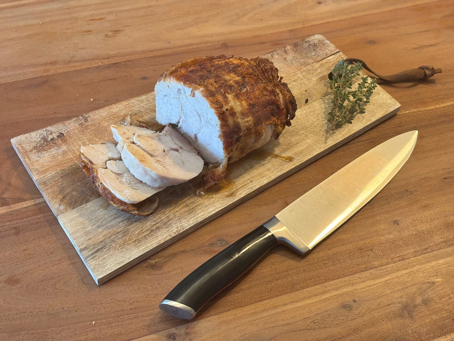 Een boerderij kiprollade, deels in plakken gesneden, ligt op een rustieke houten plank. Naast de rollade ligt een groot koksmes en een takje verse kruiden, waarschijnlijk tijm. De rollade heeft een goudbruine buitenkant en ziet er sappig uit, met duidelijk zichtbare, malse plakken kippenvlees.