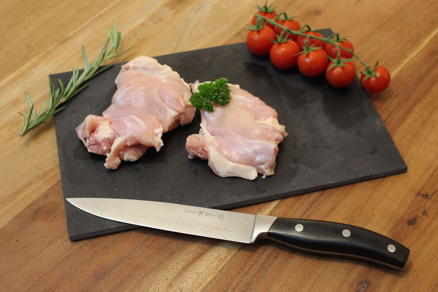 Twee kip dijen op een zwart plankje. bij de het plankje ligt een garnering van peterselie, cherrytomaten en naast de dijen ligt een takje tijm. naast het plankje ligt een koksmes.