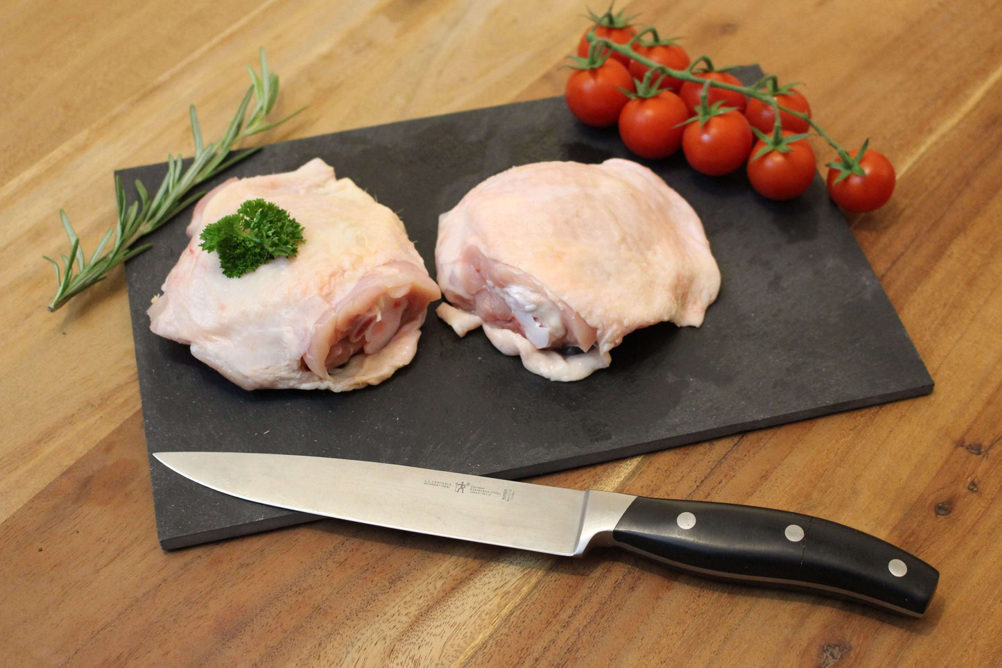 Twee kip dij karbonades op een zwart plankje. bij de het plankje ligt een garnering van peterselie, cherrytomaten en naast de dijkarbonades ligt een takje tijm. naast het plankje ligt een koksmes.