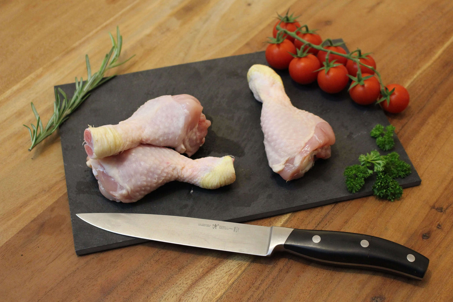 Drie kip drumsticks of poten op een zwart plankje. bij de drumsticks ligt een garnering van peterselie, cherrytomaten en naast de drumsticks ligt een takje tijm. naast het plankje ligt een koksmes.