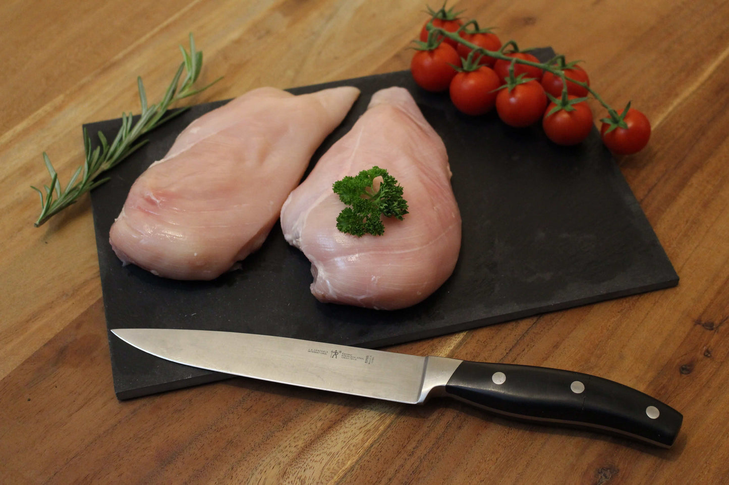 Twee sappige kipfilets op een zwart plankje. bij de het plankje ligt een garnering van peterselie, cherrytomaten en naast de filets ligt een takje tijm. naast het plankje ligt een koksmes.