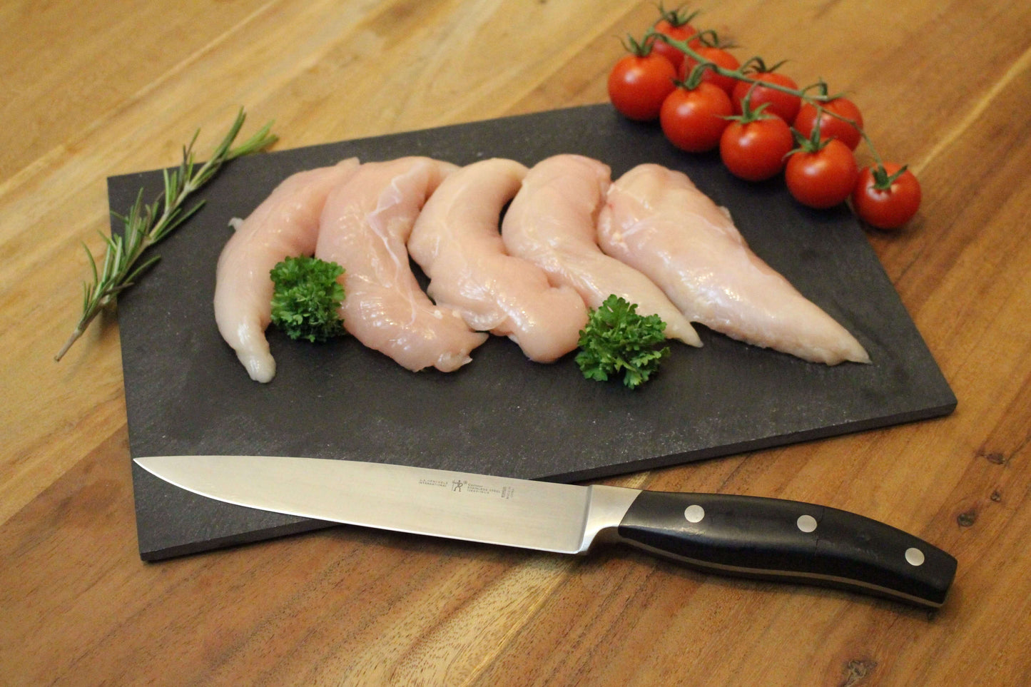 vijf kippenhaasjes op een rij op een zwart plankje. bij de haasjes ligt een garnering van peterselie, cherrytomaten en naast de haasjes ligt een takje tijm. naast het plankje ligt een koksmes.