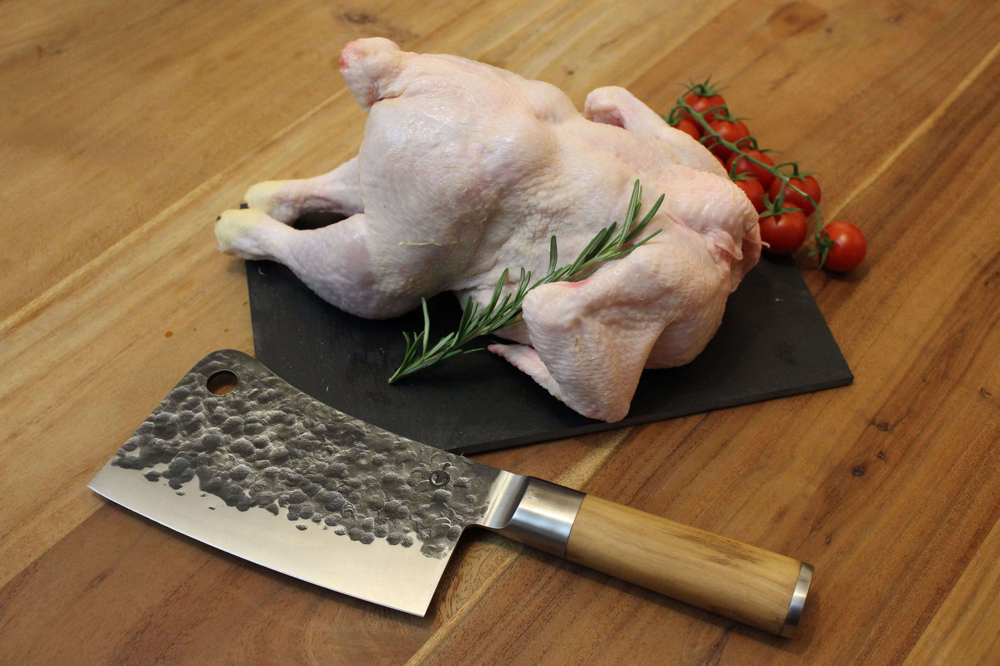 een verse hele kip op een zwart plankje. bij de kip ligt een garnering van peterselie, cherrytomaten en naast de kip ligt een takje tijm. naast het plankje ligt een koksmes.