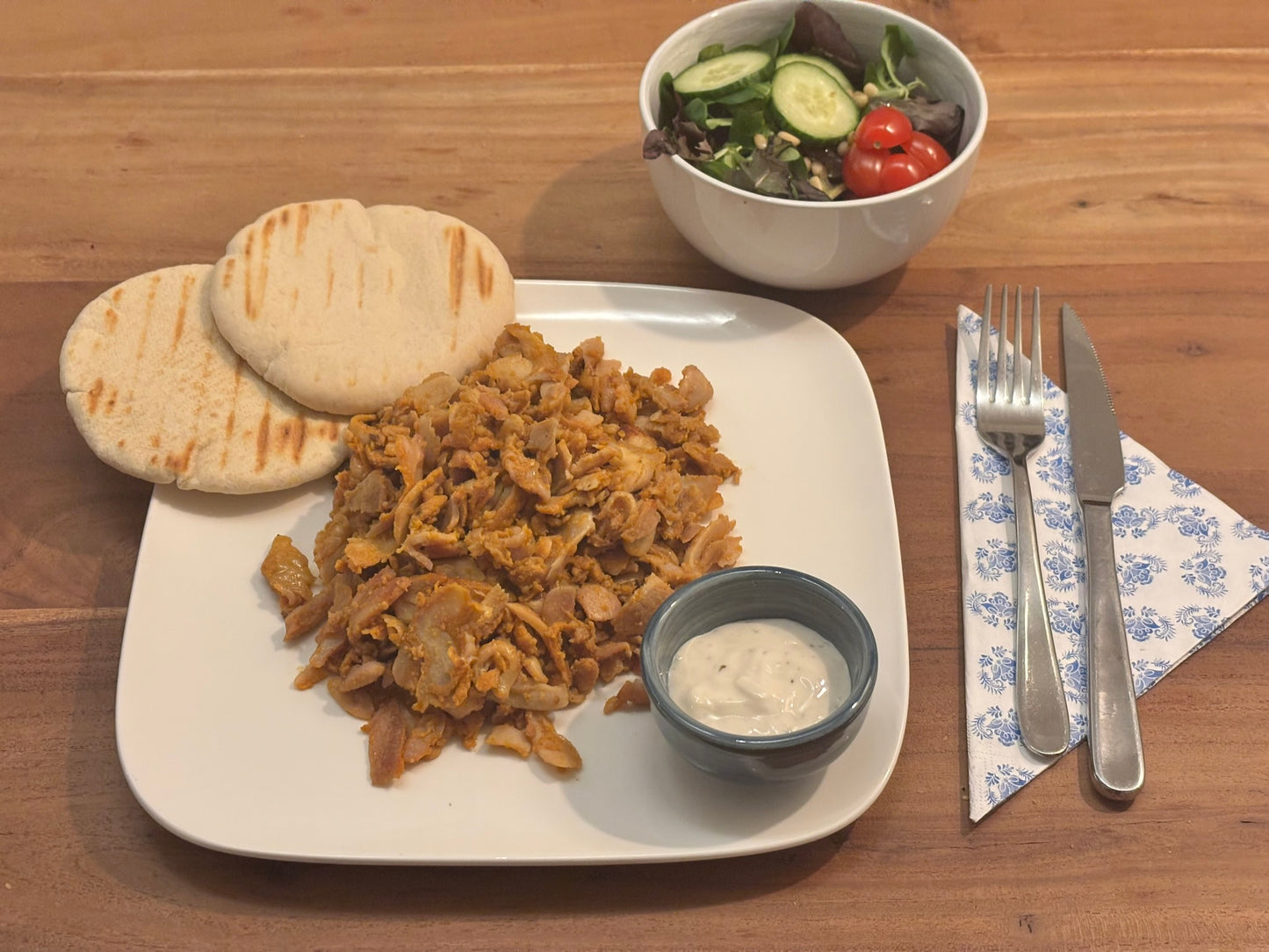 Gebakken kip kebab met een gekruid uiterlijk, geserveerd op een wit bord met twee gegrilde pita’s. Op de achtergrond een frisse salade met komkommer en cherrytomaten. Bestek ligt klaar op een servet met blauw patroon.