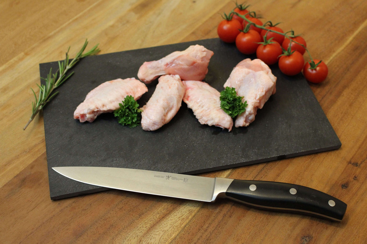 vijf kippenkluifjes op een rij op een zwart plankje. bij de kluifjes ligt een garnering van peterselie, cherrytomaten en naast de kluifjes ligt een takje tijm. naast het plankje ligt een koksmes.