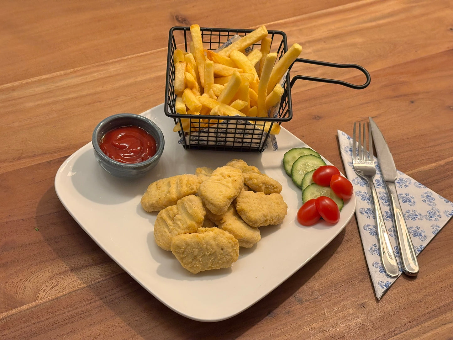 Gebakken kipnuggets met een goudbruine korst, geserveerd op een wit bord. Op de achtergrond een mandje met friet, een kommetje saus en een frisse salade met komkommer en cherrytomaten. Bestek ligt klaar op een servet met blauw patroon.