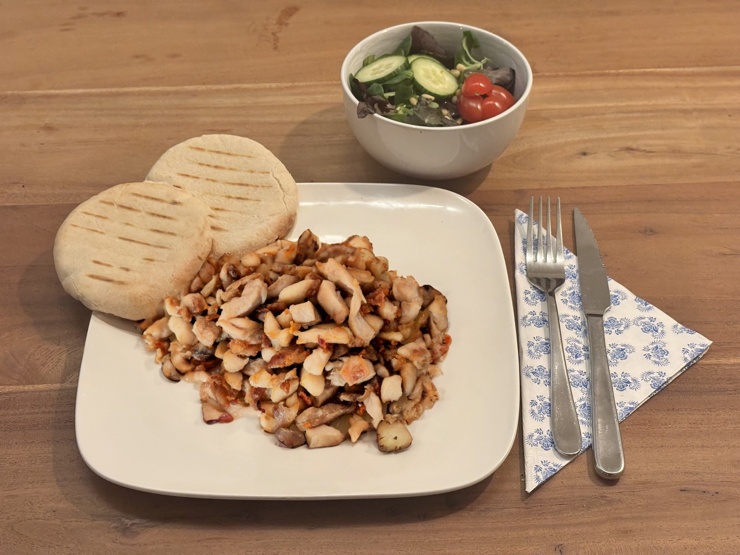 Gebakken kipreepjes met een goudbruine korst, geserveerd op een wit bord met twee gegrilde pita’s. Op de achtergrond een frisse salade met komkommer en cherrytomaten. Bestek ligt klaar op een servet met blauw patroon.