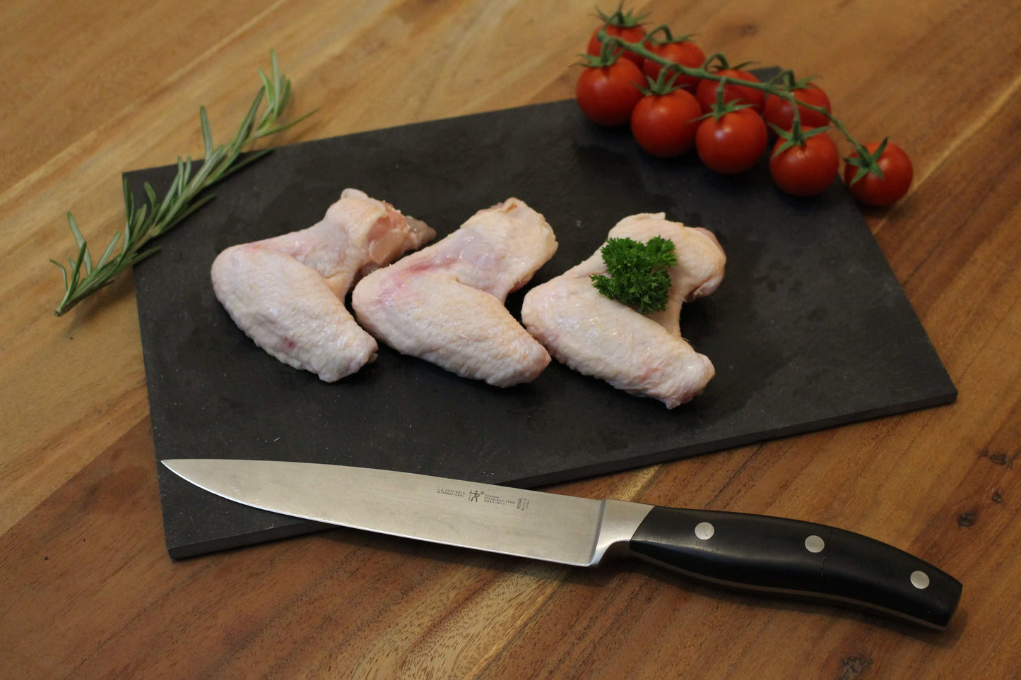 Drie kip vleugels op een zwart plankje. bij de drumsticks ligt een garnering van peterselie, cherrytomaten en naast de vleugeks ligt een takje tijm. naast het plankje ligt een koksmes.