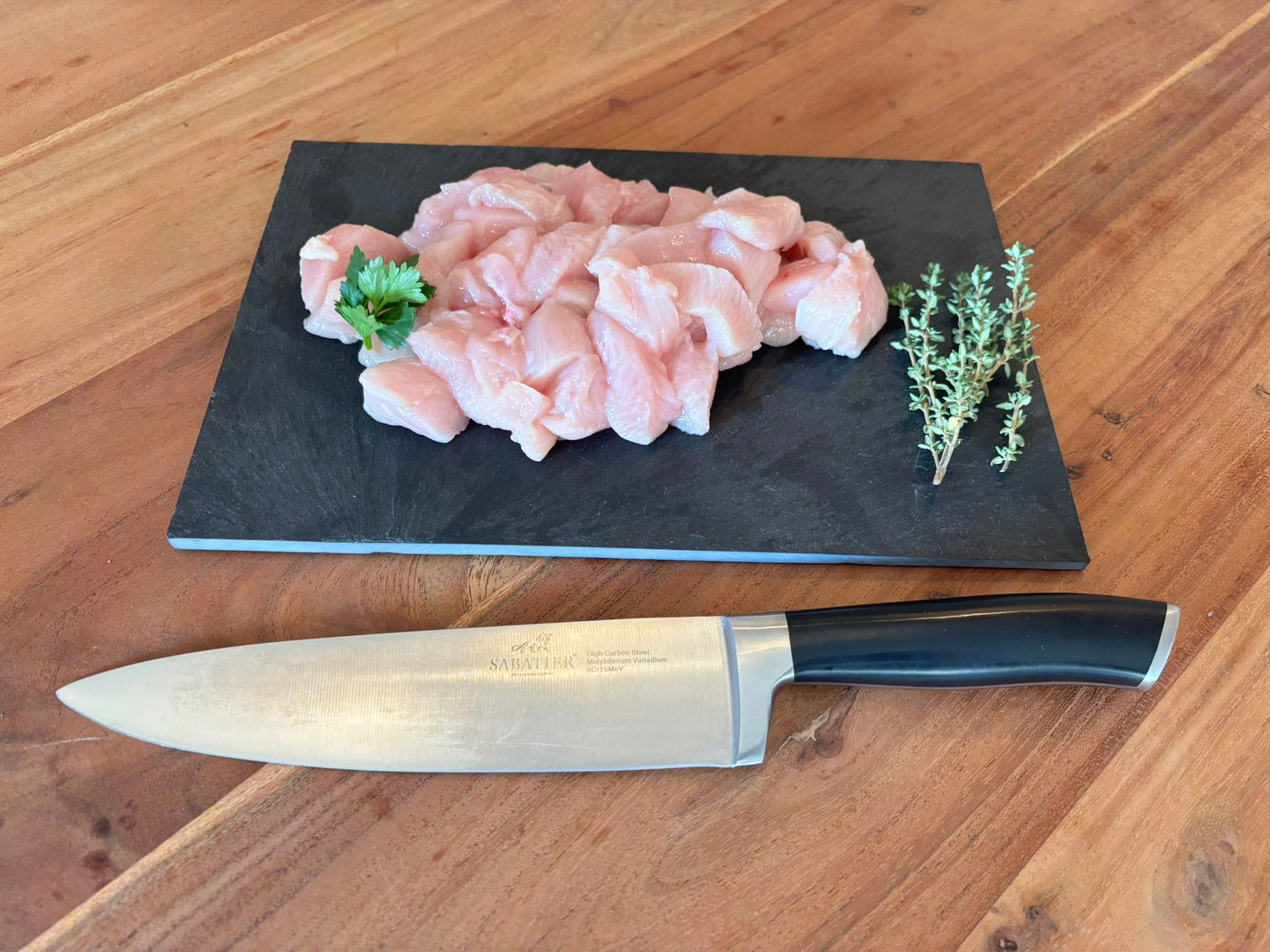 een stapel kip filet blokjes op een zwart plankje. op de blokjes ligt een garnering van peterselie en naast de blokjes ligt een takje tijm. naast het plankje ligt een koksmes.