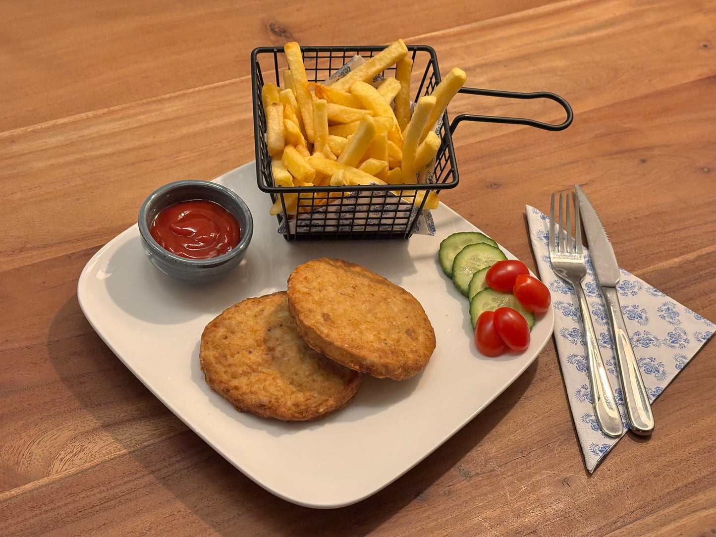 Twee knapperige kipgrillburgers, geserveerd op een wit bord. De maaltijd wordt vergezeld door een bakje friet in een metalen mandje, een kommetje saus en een frisse garnering van komkommer en cherrytomaten. Bestek ligt klaar op een blauw-wit servet.