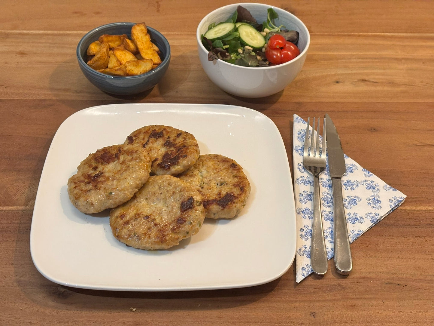 Vier knapperige kipburgers met jalepeno's en cheddar kaas, geserveerd op een wit bord. De maaltijd wordt vergezeld door aardappelpartjes in een schaal, een kommetje saus. Bestek ligt klaar op een blauw-wit servet.