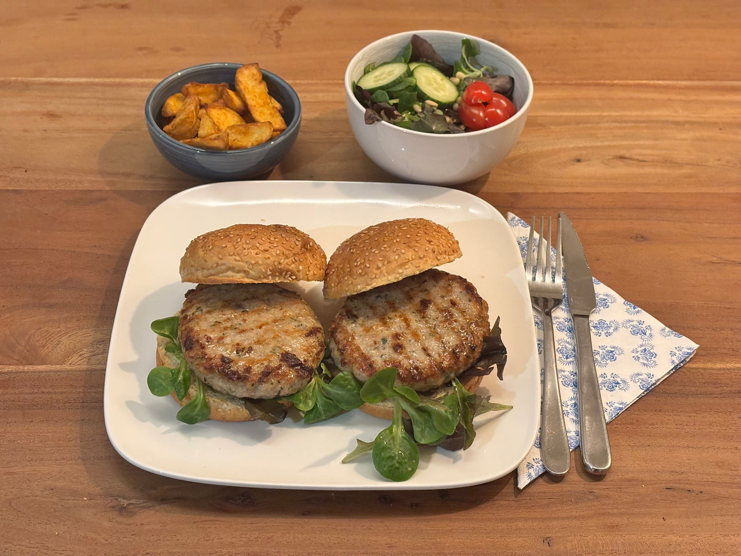 Twee knapperige kipburgers op zachte broodjes, geserveerd op een wit bord. De maaltijd wordt vergezeld door een schaaltje aardappelpartjes, een kommetje met een frisse garnering van komkommer en cherrytomaten. Bestek ligt klaar op een blauw-wit servet.