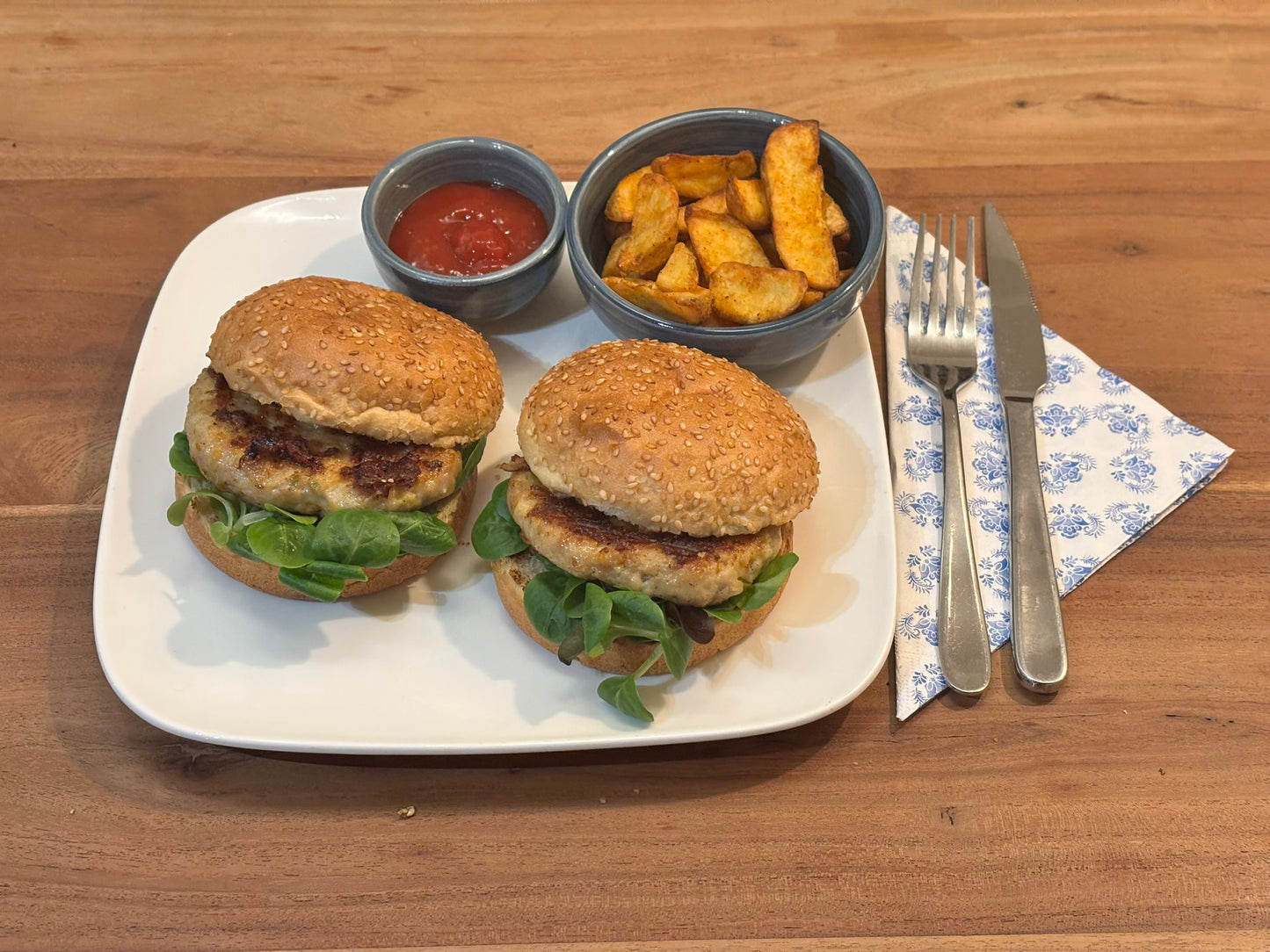 Twee knapperige kipburgers op zachte broodjes, geserveerd op een wit bord. De maaltijd wordt vergezeld door een bakje aardappel partjes in een schaal, een kommetje saus. Bestek ligt klaar op een blauw-wit servet.