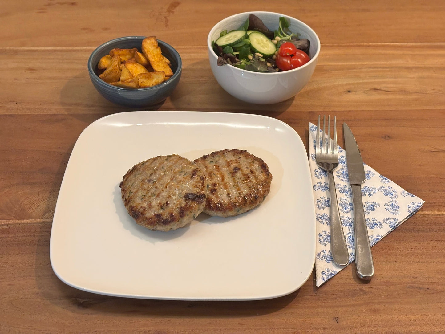 Twee knapperige kipburgers, geserveerd op een wit bord. De maaltijd wordt vergezeld door een schaaltje aardappelpartjes en een kommetje met een frisse garnering van komkommer en cherrytomaten. Bestek ligt klaar op een blauw-wit servet.