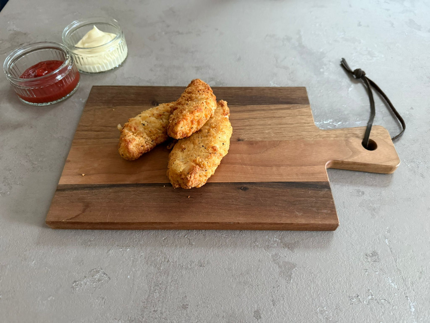 Krokante kiphaasjes geserveerd op een houten borrelplank op een leisteen ondergrond. Ze hebben een krokante paneer laag. Het product is geserveerd met een glazen schaaltje ketchup en mayonaise.