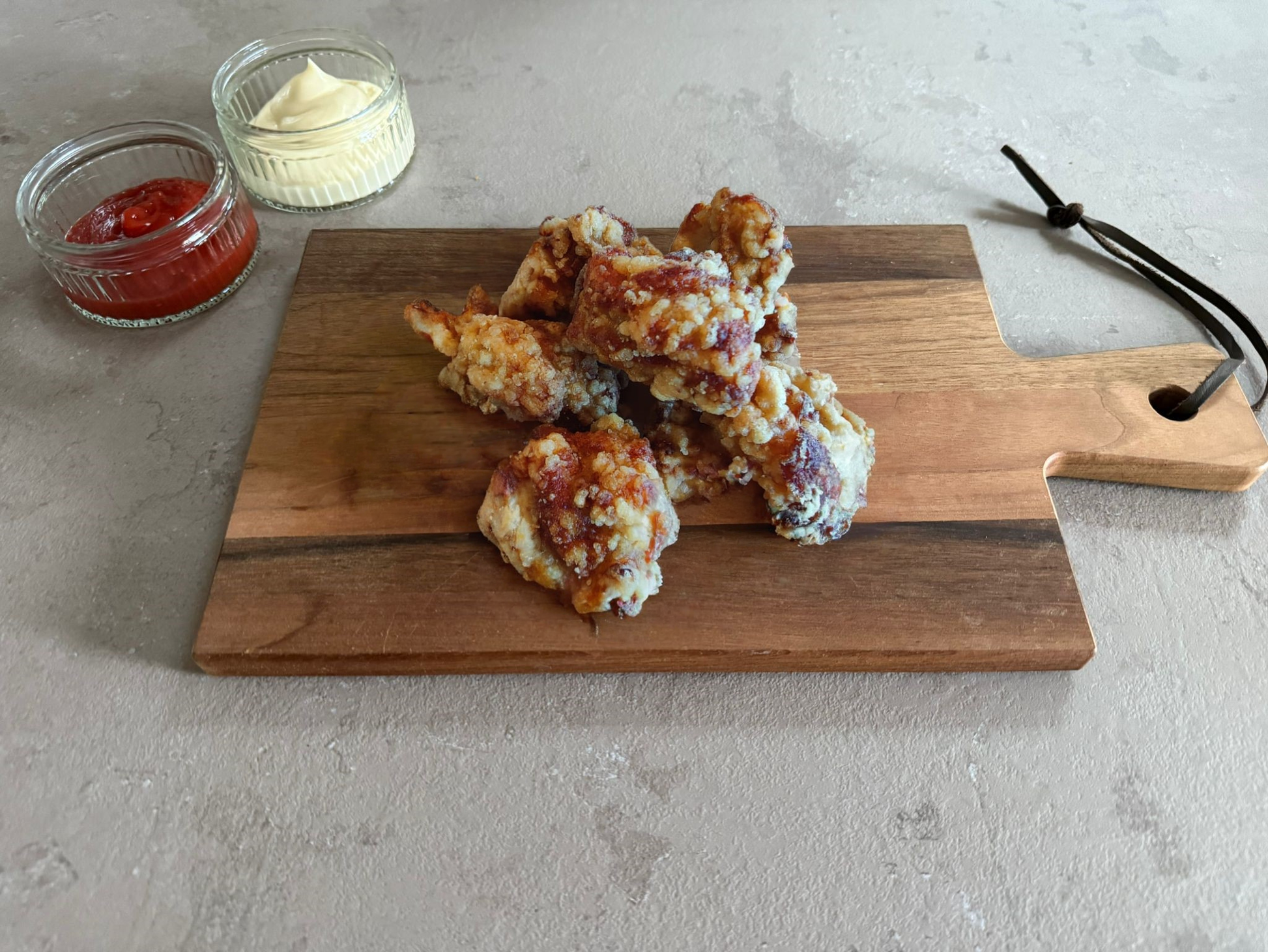 Krokante kip tatsuta chicken bites 30x30 gram geserveerd op een houten borrelplank op een leisteen ondergrond. Ze hebben een krokante paneer laag. Het product is geserveerd met een glazen schaaltje ketchup en mayonaise.
