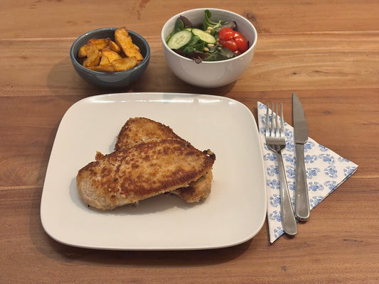 Gebakken kip schnitzels XL met een knapperige korst, omhuld met een knapperige korst. Geserveerd met krokante aardappelpartjes en een frisse salade op een houten eettafel.