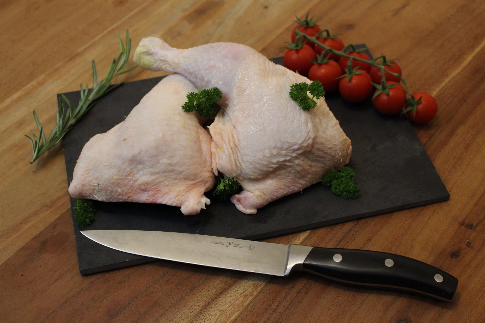 Twee kippenbouten of poten op een zwart plankje. bij de het plankje ligt een garnering van peterselie, cherrytomaten en naast de bouten ligt een takje tijm. naast het plankje ligt een koksmes.