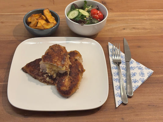 Gebakken kip cordon bleu XL met een knapperige korst, gevuld met smeltende kaas en ham. Geserveerd met krokante aardappelpartjes en een frisse salade op een houten eettafel.