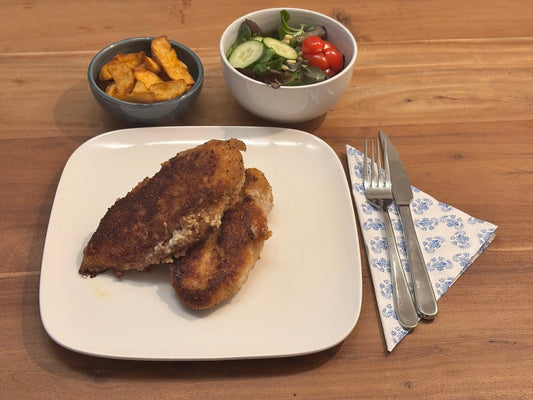 Gebakken kip cordon bleu XL met een knapperige korst, gevuld met smeltende kaas en ham. Geserveerd met krokante aardappelpartjes en een frisse salade op een houten eettafel.