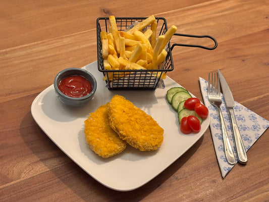 Krokant gebakken kip-schnitzels gepaneerd met met corn flakes, op een wit bord, geserveerd met een bakje friet in een metalen mandje, ketchup in een klein kommetje en een frisse garnering van komkommer en cherrytomaten. Bestek ligt klaar op een blauw-wit servet.
