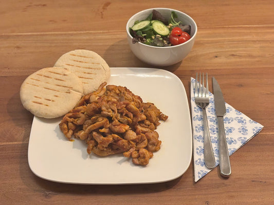 Gebakken kip shoarma met een gekruid uiterlijk, geserveerd op een wit bord met twee gegrilde pita’s. Op de achtergrond een frisse salade met komkommer en cherrytomaten. Bestek ligt klaar op een servet met blauw patroon.