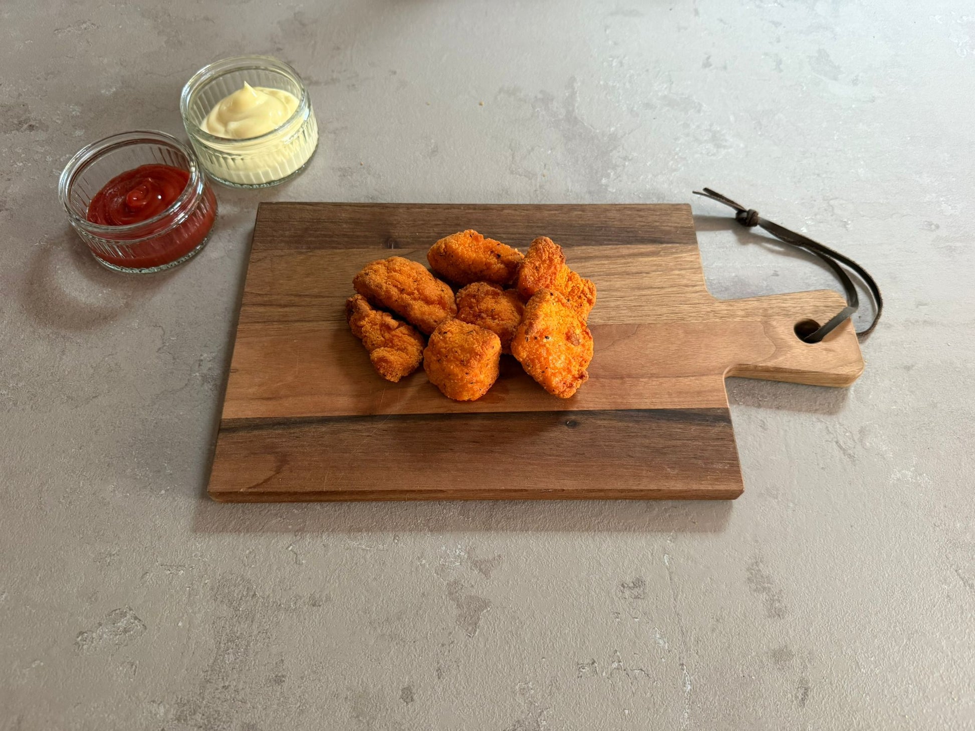 Krokante kip hot chili chunks geserveerd op een houten borrelplank op een leisteen ondergrond. Ze hebben een krokante paneer laag .Het product is geserveerd met een glazen schaaltje ketchup en mayonaise.
