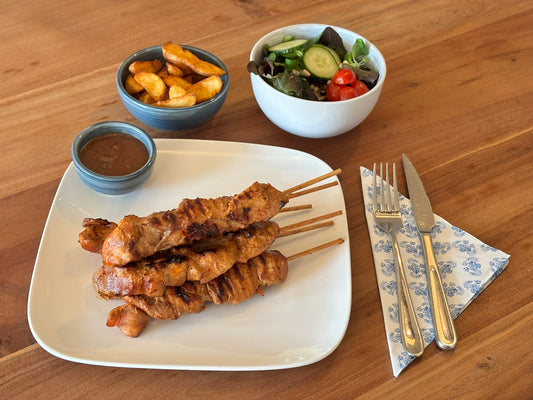 Gebakken kipdijsaté met een knapperige korst en een lekkere marinade. Geserveerd met krokante aardappelpartjes en een frisse salade op een houten eettafel.