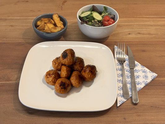 Gebakken kip gehaktballetjes met een Indische kruidenmix. Geserveerd met krokante aardappelpartjes en een frisse salade op een houten eettafel.