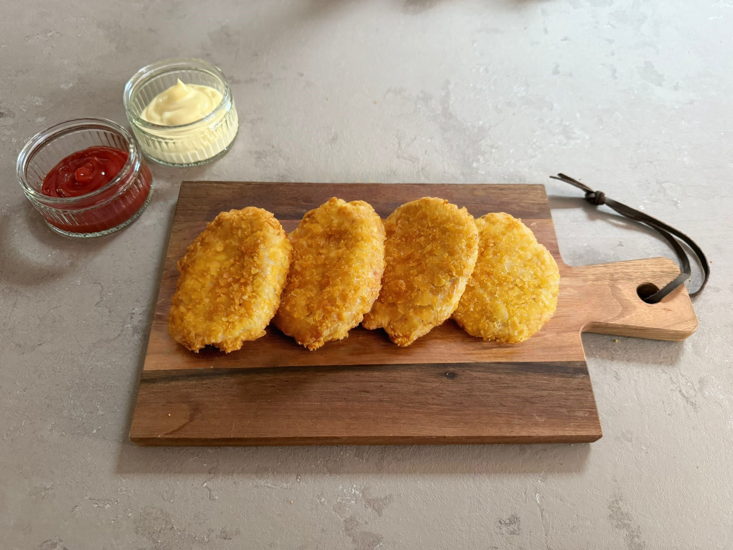 Krokante kipburgers geserveerd op een houten borrelplank op een licht grijze leisteen ondergrond. de burgers hebben een krokante paneer laag. Het product is geserveerd met een glazen schaaltje ketchup en mayonaise.