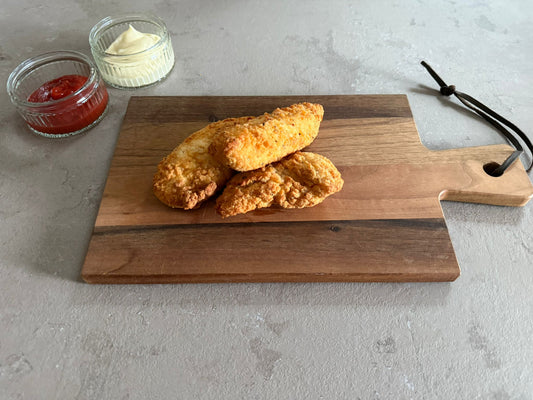 Krokante kipvingers geserveerd op een houten borrelplank op een leisteen ondergrond. Ze hebben een krokante pittige paneer laag. Het product is geserveerd met een glazen schaaltje ketchup en mayonaise.