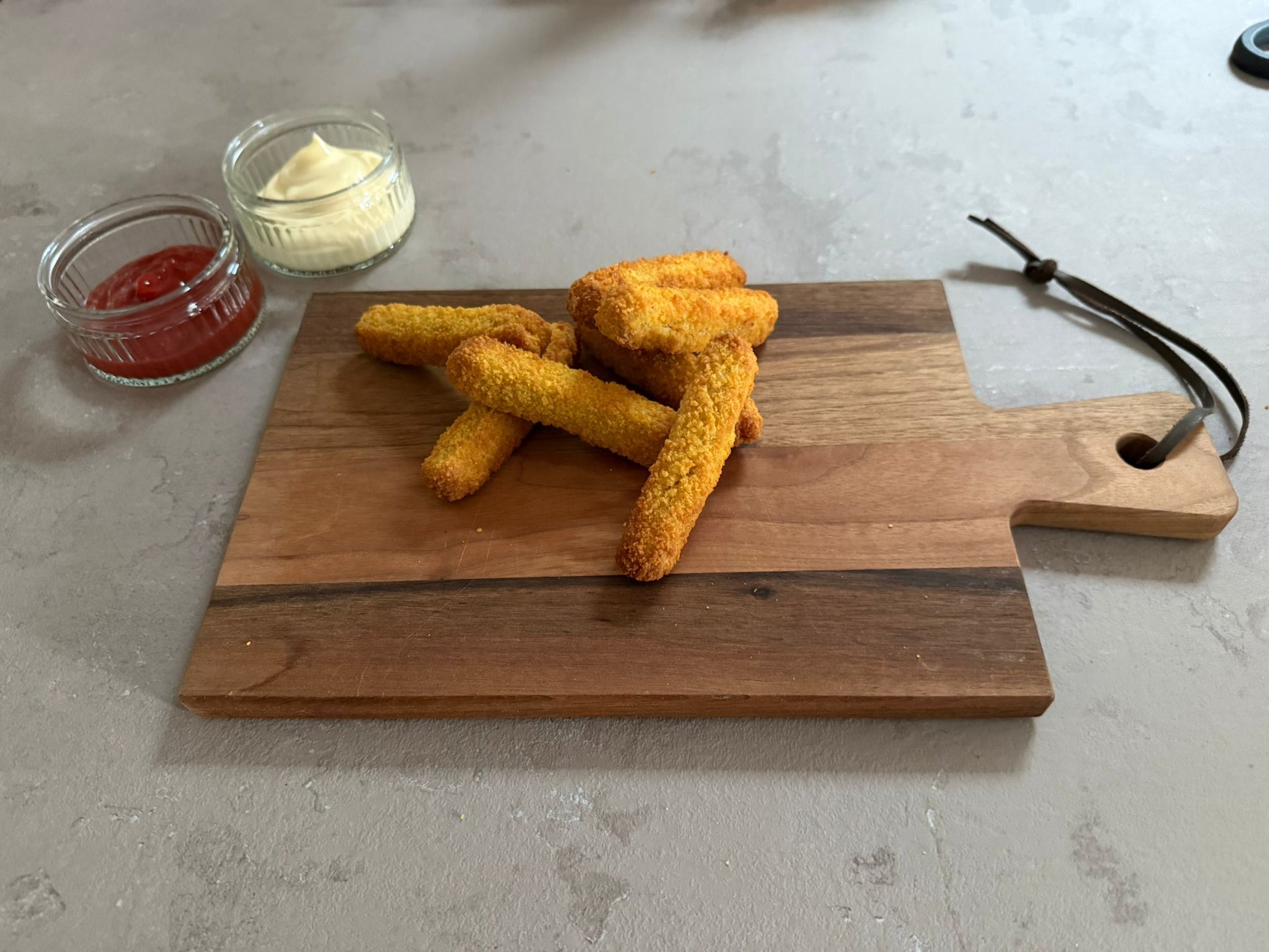 Krokante kipvingers geserveerd op een houten borrelplank op een leisteen ondergrond. Ze hebben een krokante paneer laag. Het product is geserveerd met een glazen schaaltje ketchup en mayonaise.