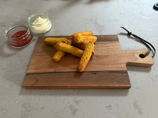 Krokante kipvingers geserveerd op een houten borrelplank op een leisteen ondergrond. Ze hebben een krokante paneer laag. Het product is geserveerd met een glazen schaaltje ketchup en mayonaise.
