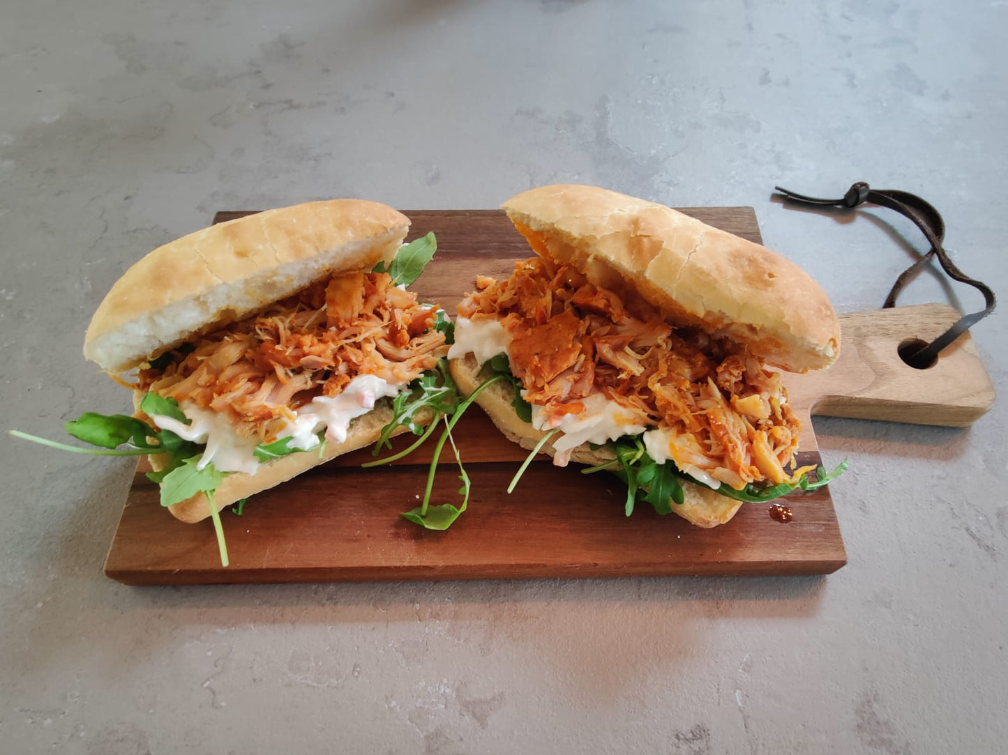 Twee broodjes gevuld met malse, sappige pulled chicken op een bedje van coleslaw en rucola. geserveerd op een rustiek houte plankje op een grijs aanrechtblad