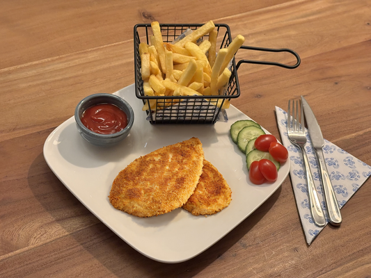 Krokant gebakken kip-schnitzels op een wit bord, geserveerd met een bakje friet in een metalen mandje, ketchup in een klein kommetje en een frisse garnering van komkommer en cherrytomaten. Bestek ligt klaar op een blauw-wit servet.